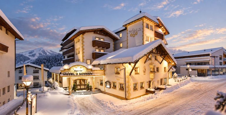 Hotel DreiSonnen in Serfaus on a winter evening: lit façade with entrance, snow-covered village street, the mountains behind in the evening light.