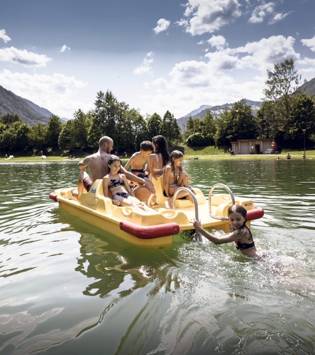 Eine Familie sitzt in einem gelben Tretboot auf dem Badesee Ried, ein Mädchen klettert aus dem Wasser an Bord.