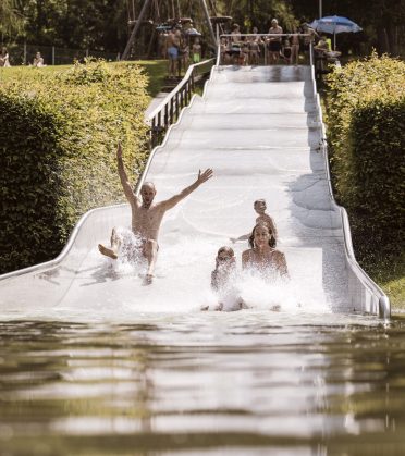 Badegäste rutschen jubelnd eine breite Wasserrutsche hinunter in den Badesee Ried.