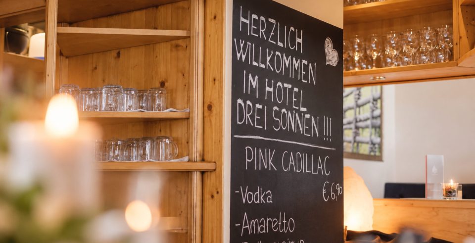 Bar im Hotel DreiSonnen: Kreidetafel mit Begrüßung und Cocktailkarte, daneben Holzregale mit Gläsern und Kerzenlicht.