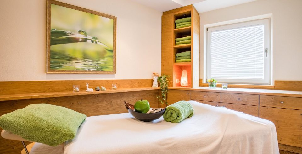 Behandlungsraum im Hotel DreiSonnen mit Massageliege, grünen Tüchern und einer Schale mit Äpfeln, an der Wand ein Bild mit einem Blatt im Wasser.