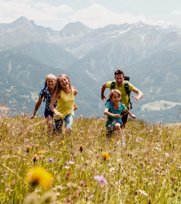 A family runs laughing through a flowering mountain meadow, the mountains in the background.
