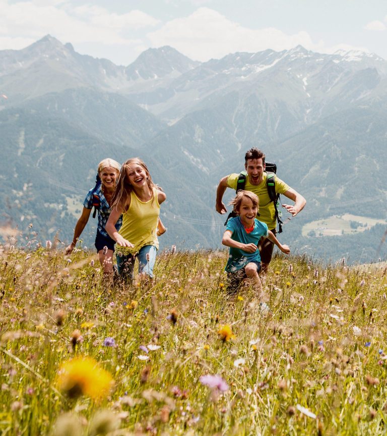 A family runs laughing through a flowering mountain meadow, the mountains in the background.