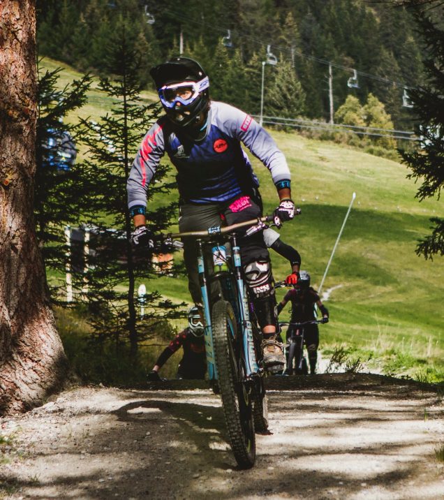 A mountain biker in helmet and goggles riding down a forest trail, more riders behind him.