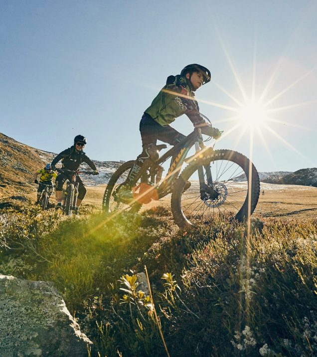 Mountainbikende fahren im Gegenlicht über eine Almwiese.
