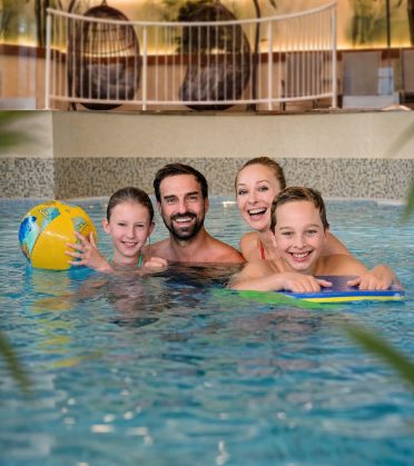 Eine Familie mit zwei Kindern lacht im Innenpool der DreiSonnen Wellnesswelt, das Mädchen hält einen Wasserball, der Junge ein Schwimmbrett.