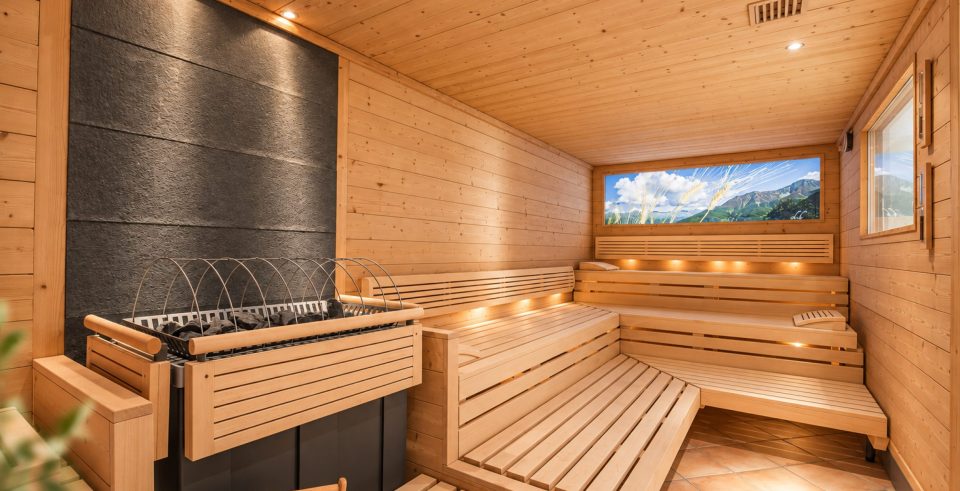 Finnische Sauna im Hotel DreiSonnen: dunkle Steinwand mit Saunaofen, Holzbänke mit warmem Licht und ein beleuchtetes Bergpanorama.