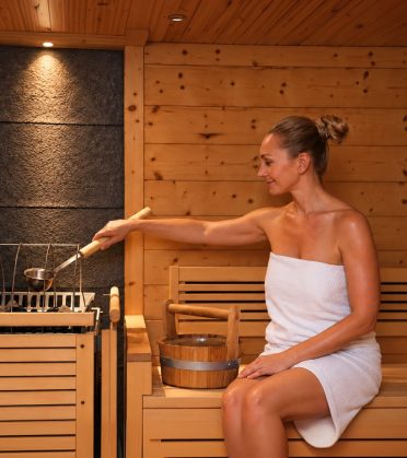 Eine Frau im Handtuch gießt in der Sauna des Hotel DreiSonnen Wasser aus der Kelle auf die Steine.