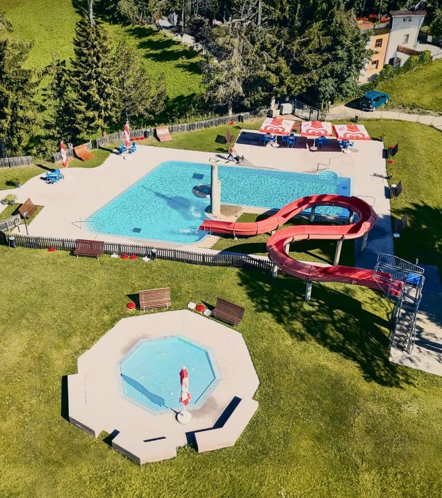 Das Freibad Serfaus von oben: Schwimmbecken mit roter Rutsche, Kinderbecken und Liegewiese zwischen Bäumen.