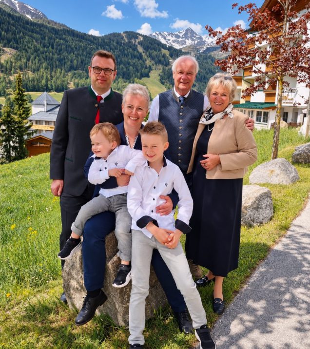 The Lechleitner host family in three generations on a meadow in Serfaus, the two boys in front, behind them Hotel DreiSonnen and the mountains.