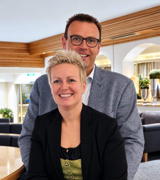 A couple at the hotel bar of Hotel DreiSonnen: she sits in front and laughs, he stands behind her in glasses and a jacket, the lobby in the background.