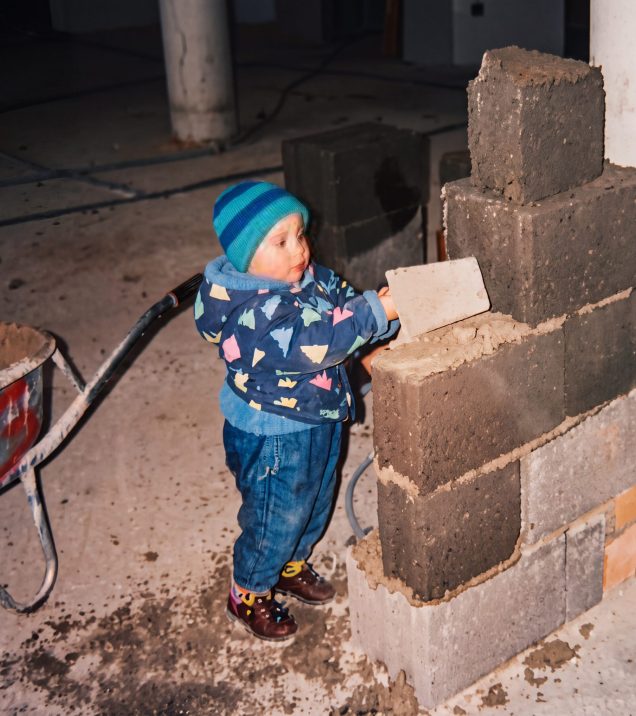 A small child in a hat places a stone on a wall of hollow blocks at a building site, a wheelbarrow beside them.