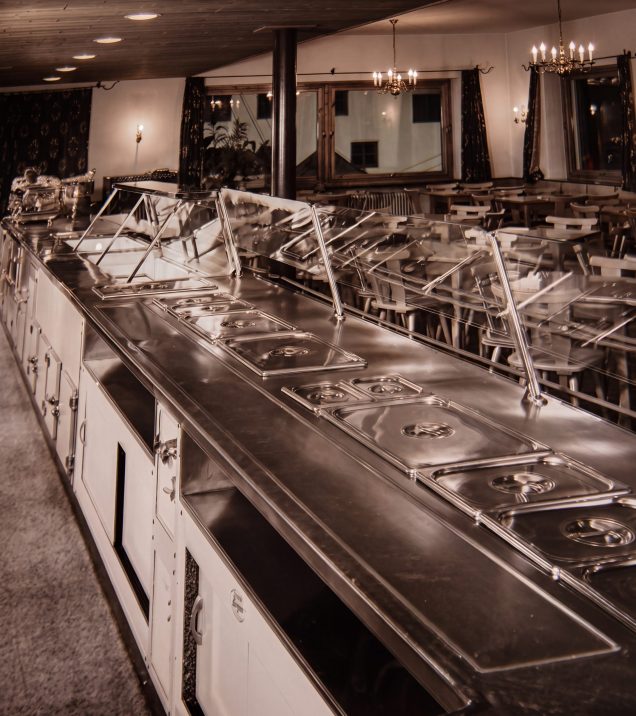 Historical photo of a long buffet counter with warming trays, behind it a dining room with tables and chandeliers.