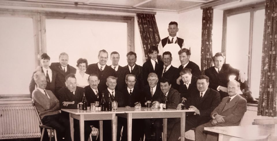 Historic black-and-white photo: a group of men in suits at long tables in a parlour with large windows, behind them a man in a white jacket.