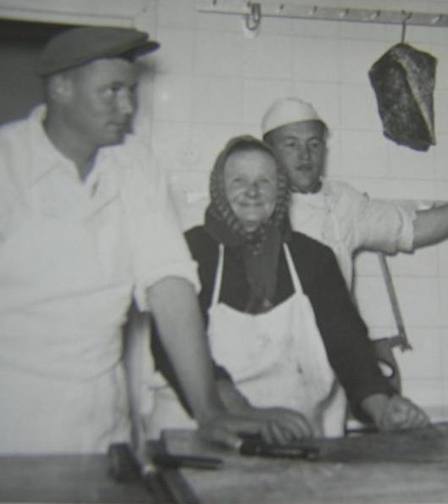 Historic black-and-white photo from the butcher's shop: two men in white work clothes and a woman with a headscarf and apron at the work table, meat hanging from a hook on the wall.