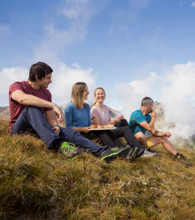 Four walkers sit in the grass on the mountain taking a rest, clouds drifting past behind them.