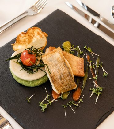 Ein Fischgang mit Gemüse und Kresse auf einem grauen Teller, von oben fotografiert, daneben Besteck und ein Glas im warmen Abendlicht.