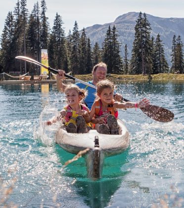A father paddles a canoe across a mountain lake with two girls in life jackets.
