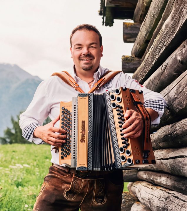 Ingo Stecher with his Styrian accordion in front of a wooden hut, behind him a mountain meadow and the mountains.