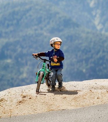 A small boy in a helmet pushes his children's bike over a hump in the bike park.