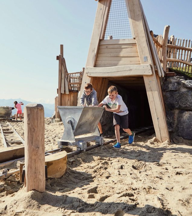 Two boys pushing a mine cart out of a wooden tunnel through the sand.