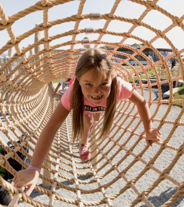 A girl climbs laughing through a rope-net tunnel.