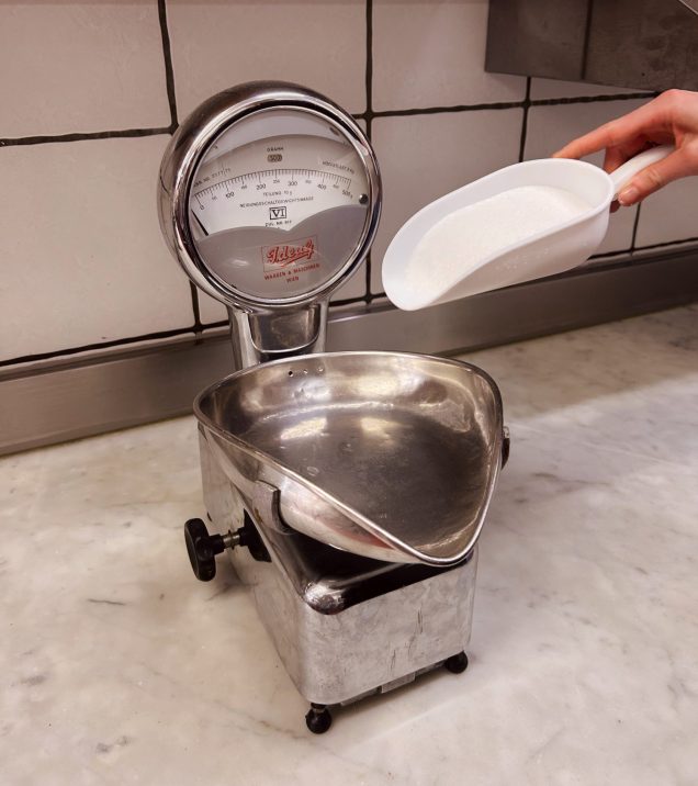 A hand scooping a white ingredient into an old metal kitchen scale.