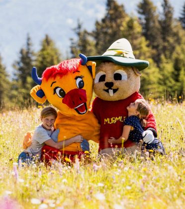 Two children hug the mascot Murmli and a second figure in a flower meadow.