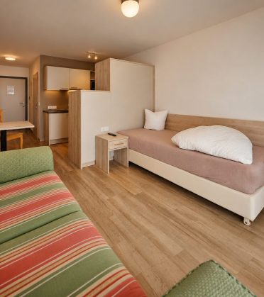 Room in the staff house of Hotel DreiSonnen with a bed, a striped sofa and a kitchenette in the background.