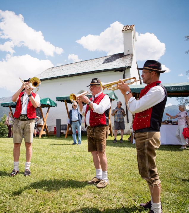 Three musicians in traditional dress play trumpets on a meadow in front of a small church, guests under parasols behind them.