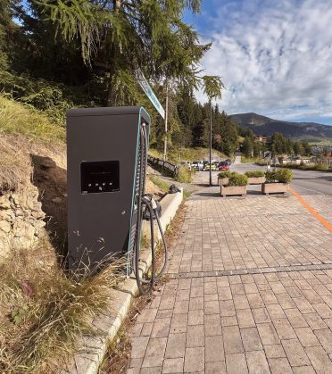 A public EV charging point at the edge of a paved footpath in Serfaus, behind it trees, a car park and the mountains.