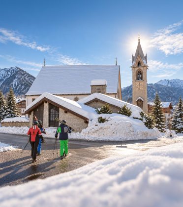 Skifahrer mit geschulterten Ski gehen an einem sonnigen Wintertag durch den verschneiten Ortskern von Serfaus, dahinter die Kirche mit ihrem Turm und die Berge.