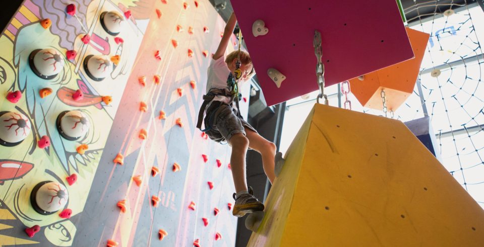 A boy climbs a colourful climbing wall next to a large yellow climbing cube.