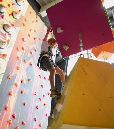 A boy climbs a colourful climbing wall next to a large yellow climbing cube.