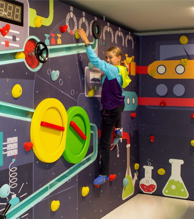 A girl climbs a play wall with wheels, tubes and laboratory jars.