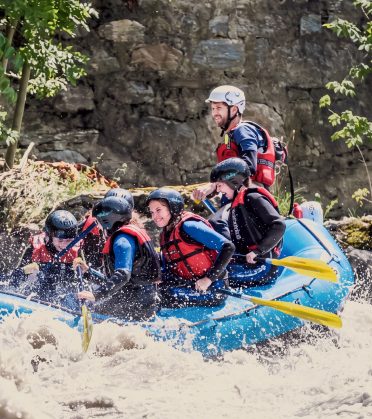 A group in helmets and life jackets paddles a blue raft across the river.