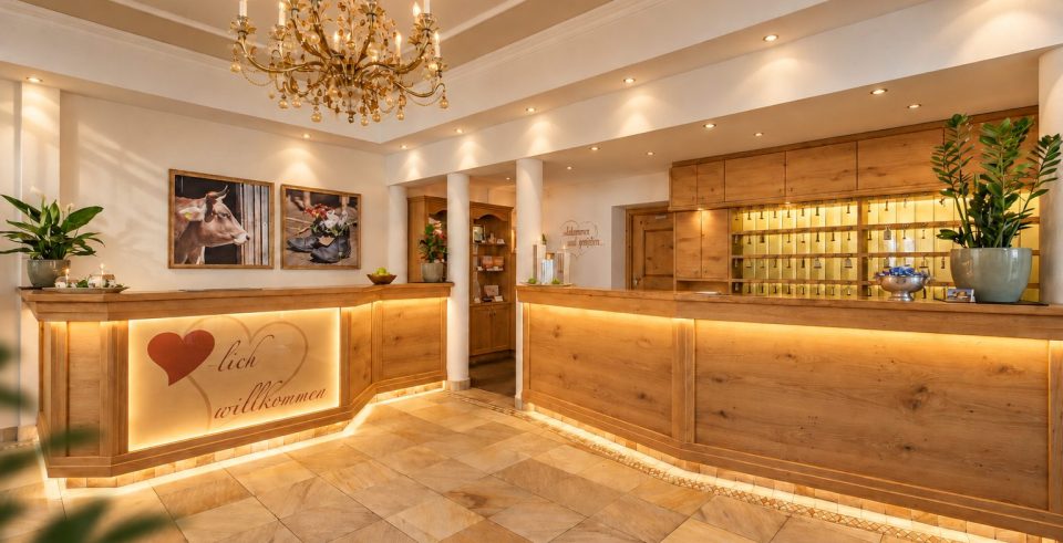 The reception of Hotel DreiSonnen in Serfaus with an illuminated counter of light wood, a chandelier and a key board