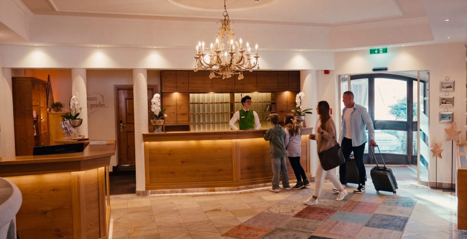 The reception of Hotel DreiSonnen with a chandelier and an illuminated wooden counter, a member of staff welcoming a family with suitcases.