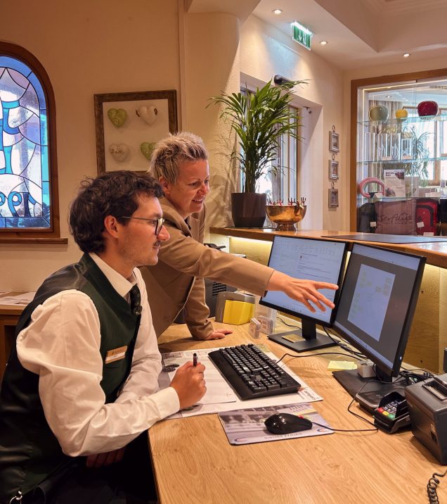 A staff member shows a young colleague something on the screen at the reception of Hotel DreiSonnen.