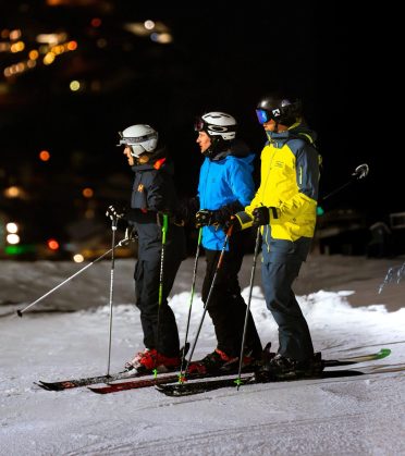 Three skiers on the floodlit piste in the evening, the lights of the village behind them.