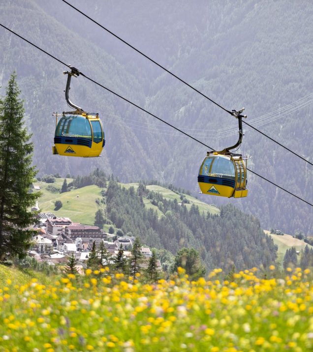 Two yellow gondolas of a cable car float above a meadow with yellow flowers, in the valley a village between forest and alpine meadows.