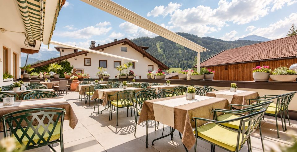 Sonnenterrasse des Hotel DreiSonnen im Sommer: gedeckte Tische mit Blumen und grünen Stühlen unter der Markise, dahinter Berge und blauer Himmel.