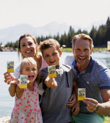 A family at a mountain lake holds their Super.Sommer.Cards up to the camera, laughing.