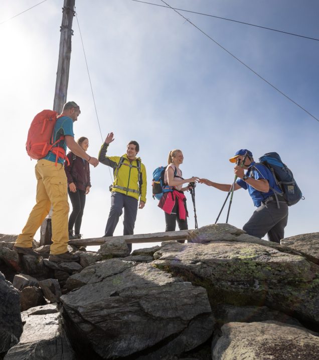 A group of hikers with rucksacks and poles at the summit cross on a rocky peak, two of them shaking hands.