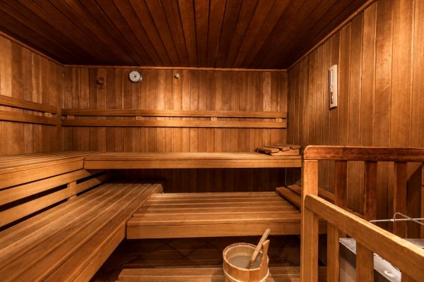 Sauna mit dunkler Holzverkleidung und Holzbänken auf mehreren Stufen, vorne ein Holzkübel mit Schöpfkelle.