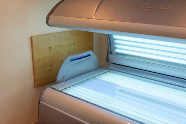 Geöffnete Solarium-Liege mit leuchtenden Röhren in einem hellen Raum, dahinter eine Holzwand.