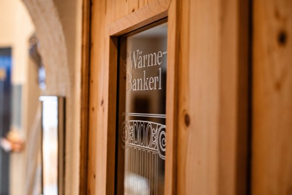 Glastür aus Holz mit der eingravierten Aufschrift Wärme Bankerl, dahinter unscharf der Gang im Wellnessbereich.