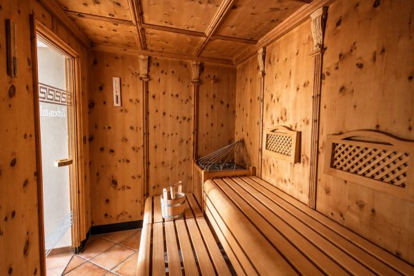 Innenansicht der Zirbensauna mit Wänden und Bänken aus hellem Zirbenholz, geschnitzten Gittern und einer Glastür links.