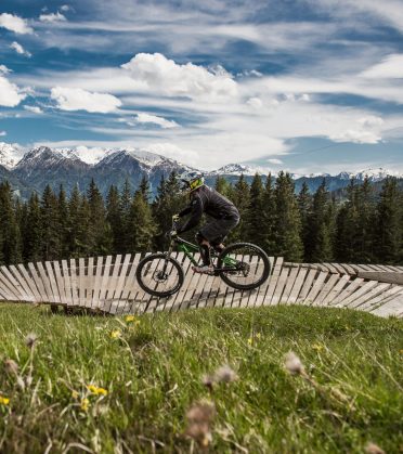 A female mountain biker riding over a curved wooden bridge at the edge of the forest.