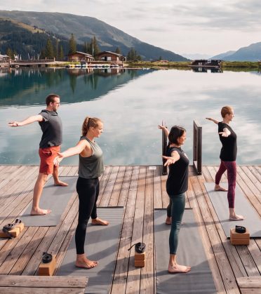 A yoga group practising on mats on the jetty of the Högsee, the lake and the mountains behind them.